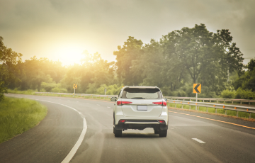 White SUV driving down the road with sun and trees in the distance