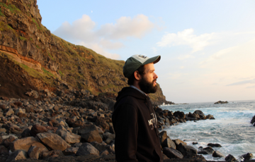 Bearded man wearing black hoodie and hat looking at the ocean